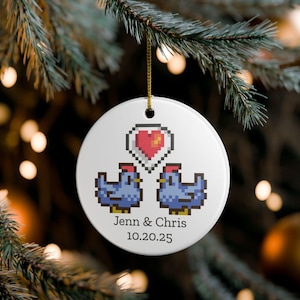 Peut inclure: Une décoration de Noël en céramique blanche avec un design pixélisé. Deux poulets bleus se font face sous un cœur rouge pixélisé. Les noms "Jenn & Chris" et la date "10.20.25" sont imprimés en dessous. Une ficelle dorée est attachée pour l'accrocher.