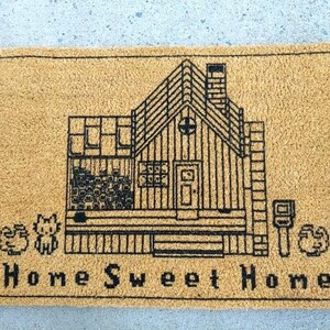 Puede incluir: Una alfombra rectangular con un fondo beige y un dibujo lineal negro de una casa. La casa tiene una chimenea, ventanas y una puerta. Las palabras "Home Sweet Home" est&aacute;n impresas en negro en la parte inferior. Tambi&eacute;n hay dos peque&ntilde;as im&aacute;genes decorativas en la alfombra.