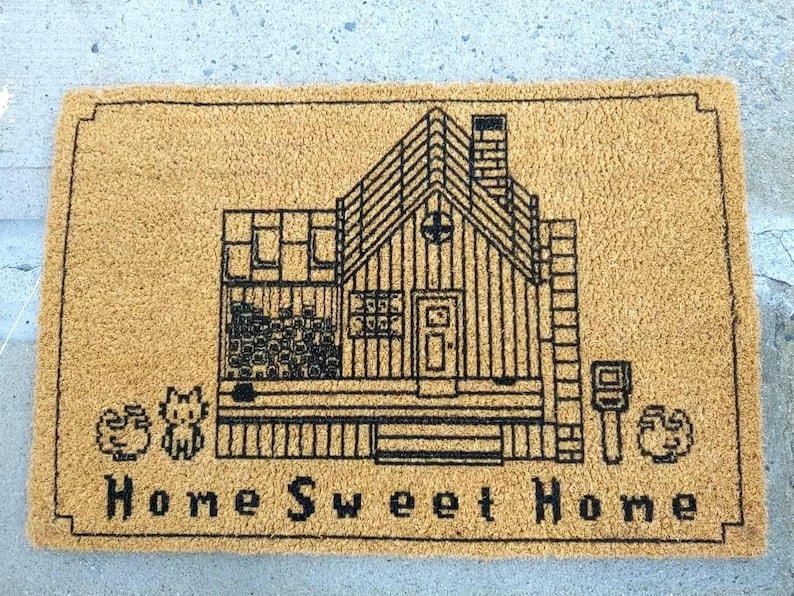 Puede incluir: Una alfombra rectangular con un fondo beige y un dibujo lineal negro de una casa. La casa tiene una chimenea, ventanas y una puerta. Las palabras "Home Sweet Home" est&aacute;n impresas en negro en la parte inferior. Tambi&eacute;n hay dos peque&ntilde;as im&aacute;genes decorativas en la alfombra.