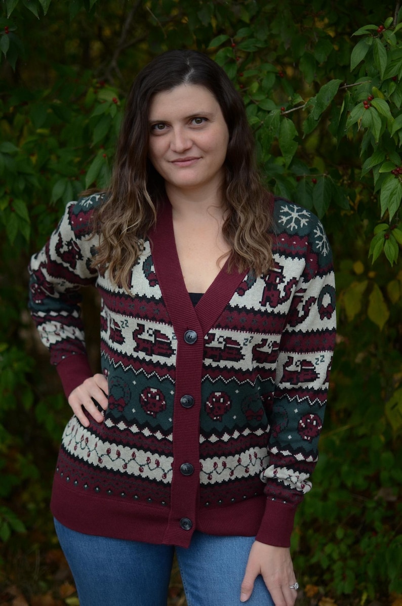 May include: A burgundy cardigan sweater with a festive pattern featuring snowflakes, reindeer, and other holiday motifs. The sweater has a V-neck, button closure, and ribbed cuffs. The background is a natural green.