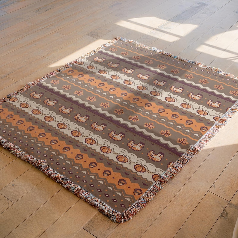 May include: A brown and orange throw blanket with a repeating pattern of pumpkins, chickens, and geometric shapes. The blanket has a fringed edge and is made of a soft, woven material. The design is reminiscent of autumn.