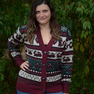 May include: A burgundy, green, and cream-colored cardigan sweater with a V-neck and button closure. The sweater features a festive pattern with snowflakes, trucks, and other holiday-themed designs. The cuffs and trim are burgundy.