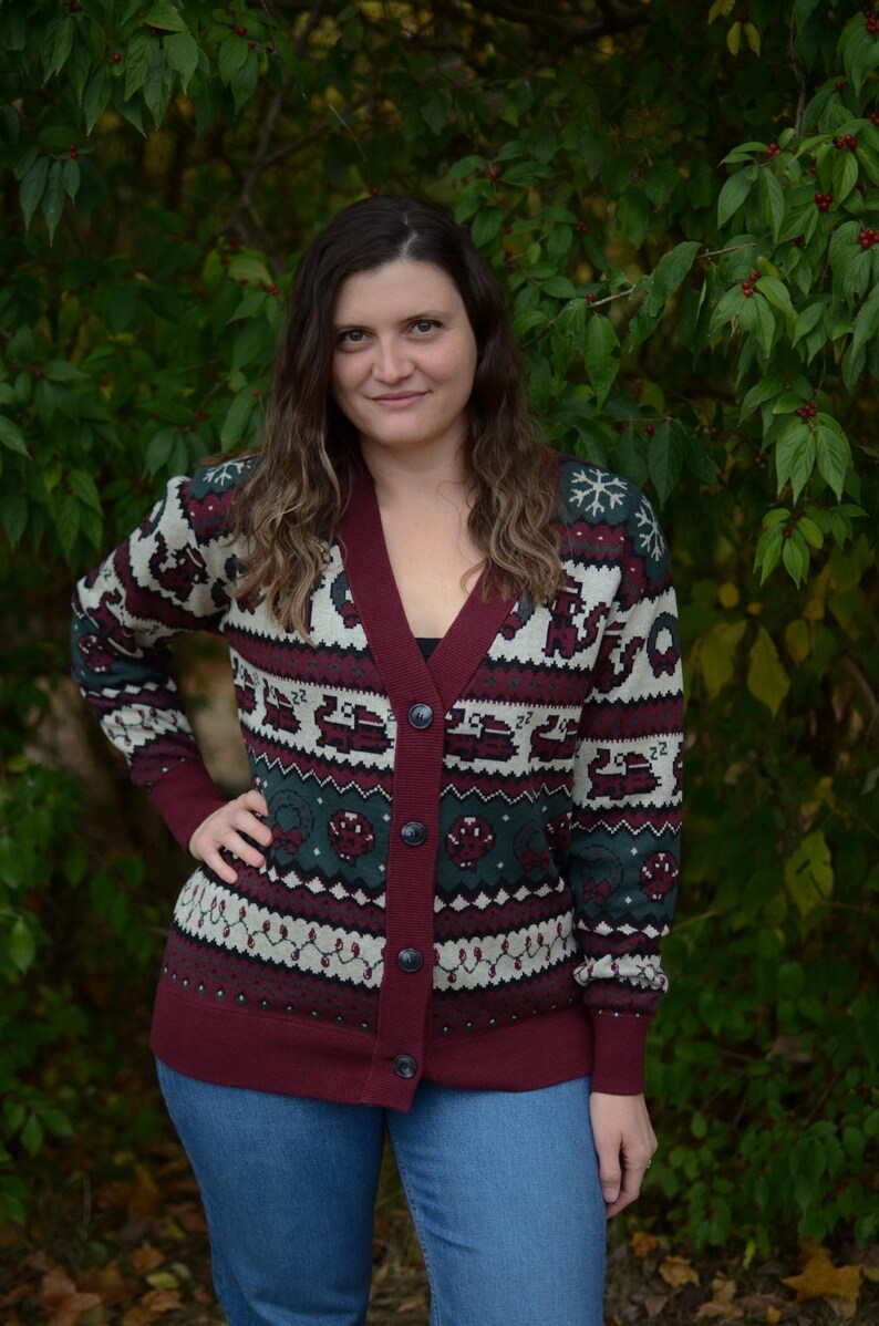 May include: A burgundy, green, and cream-colored cardigan sweater with a V-neck and button closure. The sweater features a festive pattern with snowflakes, trucks, and other holiday-themed designs. The cuffs and trim are burgundy.
