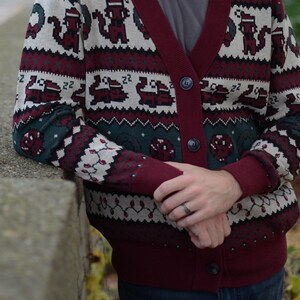 May include: Burgundy and green Christmas-themed cardigan sweater with a button-up front. The sweater features a pattern of pixelated animals, snowflakes, and other holiday designs. The cuffs and trim are a solid burgundy color.