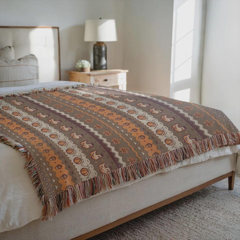 May include: A woven throw blanket with a brown background and a repeating pattern of pumpkins, horses, and geometric shapes in orange, white, and burgundy. The blanket has a fringed edge and is draped over a bed.