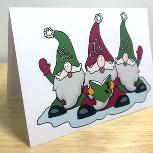 May include: Three gnomes singing Christmas carols. The gnome in the middle is wearing a red hat and holding a book with music notes. The gnomes on either side are wearing green hats. The gnome on the left is waving. The text on the hats reads "Fa", "La", and "La".