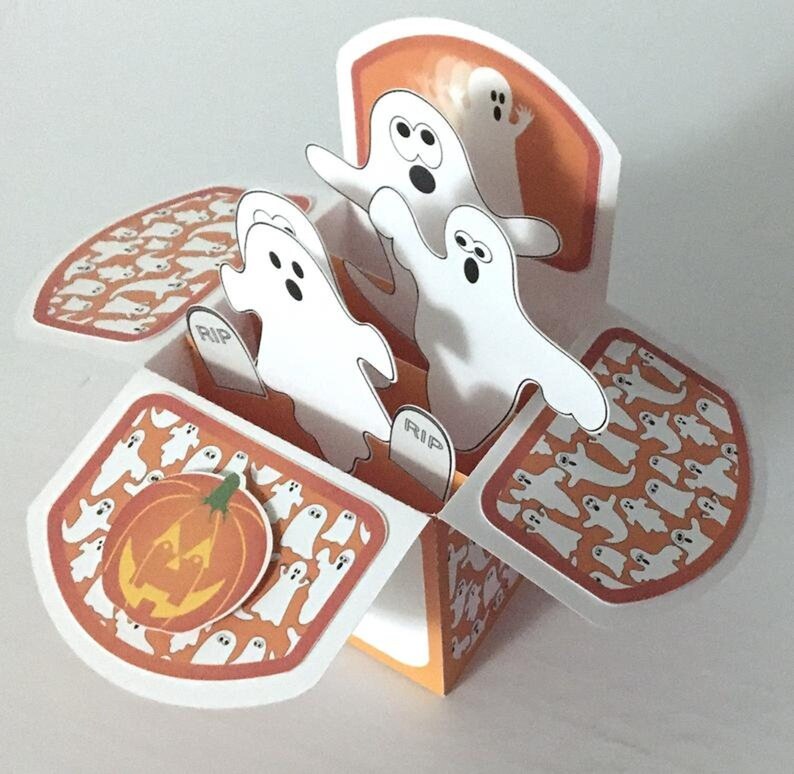 May include: A pop-up card with a Halloween theme. The card features a pumpkin and three ghosts with the word "RIP" on the tombstones. The background is orange with a pattern of white ghosts.