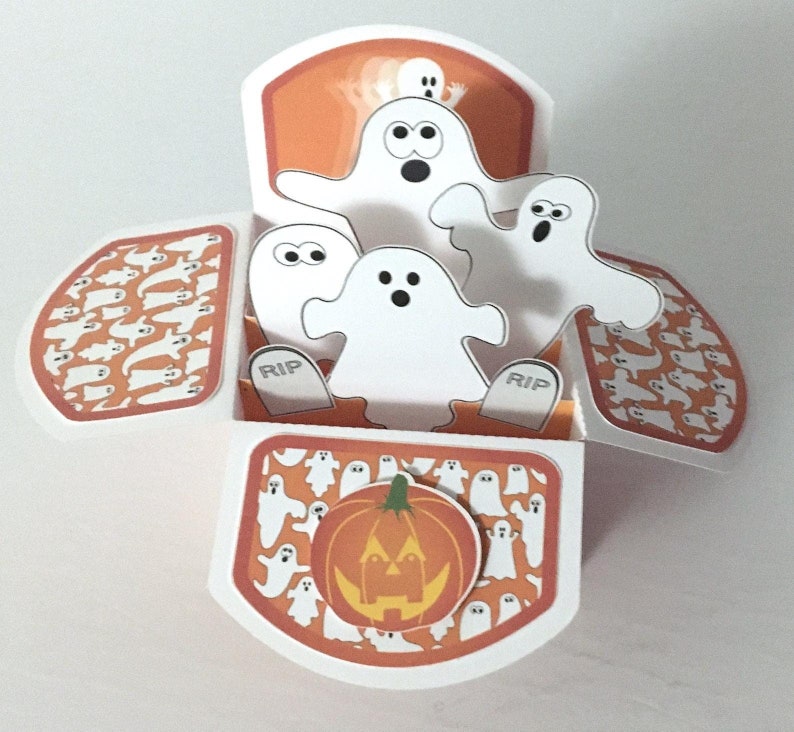 May include: A pop-up card with a Halloween theme. The card features a jack-o'-lantern and several ghosts. The ghosts are white and have black eyes and mouths. The card is decorated with a pattern of ghosts on an orange background.