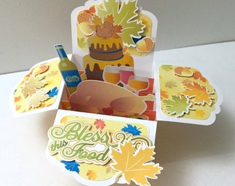 May include: A pop-up card with a Thanksgiving theme. The card features a turkey, a cake, wine glasses, and a bottle of wine. The card is decorated with fall leaves and the words "Bless this Food".