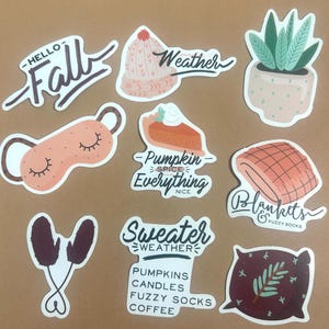 Peut inclure: Un ensemble de neuf autocollants sur le thème de l'automne avec des illustrations d'un pull confortable, d'une tarte à la citrouille, d'un masque de sommeil, d'une plante en pot, d'une paire de moufles, d'une couverture, d'un oreiller et de deux autocollants textuels qui disent "Hello Fall" et "Pumpkin Spice Everything Nice".