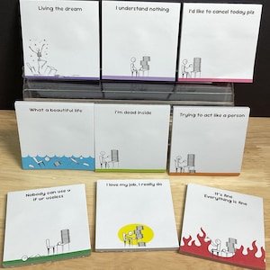 Peut inclure: Un ensemble de post-it carrés avec des phrases humoristiques et des illustrations de bonhommes allumettes. Les phrases incluent "Living the dream", "I understand nothing" et "I'd like to cancel today plz". Les notes ont des bordures colorées et sont disposées sur une surface en bois.
