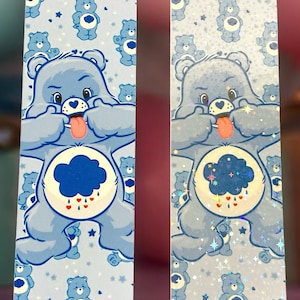 Peut inclure: Deux marque-pages rectangulaires avec un motif d'ours bleu clair. L'image principale est un Care Bear tirant la langue. Le fond est bleu clair avec des illustrations de petits Care Bears et des étoiles.