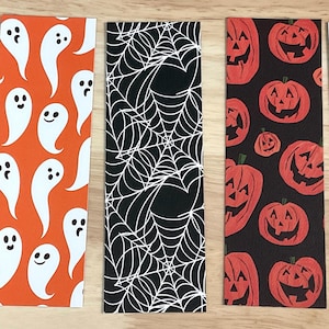 May include: Five Halloween-themed bookmarks. Designs include bats, ghosts, spiderwebs, jack-o'-lanterns, and skulls. The bookmarks are rectangular and feature various colours such as orange, black, and gold.
