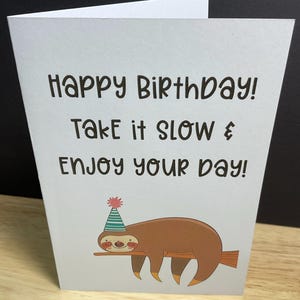 May include: A folded birthday card with the text "Happy Birthday! Take it slow & Enjoy your day!" features a cartoon sloth wearing a party hat and resting on a branch. The card is white with black text and a brown sloth illustration.