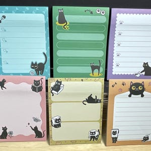 May include: Six cat-themed sticky note pads in various colours. Each pad features a different design, with illustrations of black cats in different poses. The pads have lined pages for writing notes and reminders.