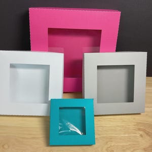 May include: A collection of colorful, square gift boxes with clear window cutouts. The boxes are in various sizes and colors, including pink, white, gray, and teal. The boxes are arranged on a wooden surface.