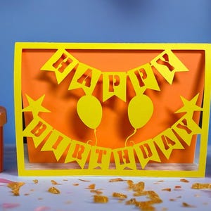May include: A yellow paper cut-out birthday card with the words "HAPPY BIRTHDAY" and two balloons. The card is set against an orange background and framed in yellow. Pink gift boxes with ribbons are on either side.