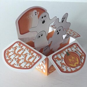 May include: A pop-up card with a Halloween theme. The card is open and features a pumpkin with a jack-o'-lantern face and a group of ghosts popping up from the inside. The card is decorated with a pattern of ghosts on an orange background.