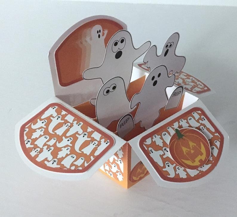 May include: A pop-up card with a Halloween theme. The card is open and features a pumpkin with a jack-o'-lantern face and a group of ghosts popping up from the inside. The card is decorated with a pattern of ghosts on an orange background.