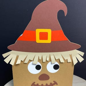 May include: A square tan box decorated as a scarecrow face with a brown hat featuring an orange band and yellow buckle. The face has two large white eyes with black pupils, a brown nose, and a stitched mouth. Beige paper fringe mimics straw hair.
