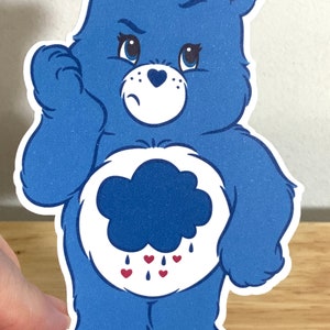 May include: A blue Care Bear with a grumpy expression and a dark blue cloud with red hearts raining from it on its belly. The bear is standing with its arms crossed.