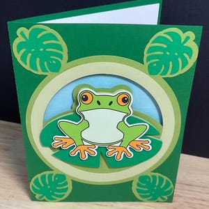 May include: A folded greeting card featuring a layered paper design with a central green frog sitting on a lily pad. The card has a circular frame, a light blue background, and four decorative monstera leaves in the corners on a dark green base.