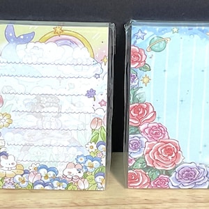May include: Four rectangular memo pads wrapped in clear plastic. The first features hot air balloons and tulips. The second shows a rainbow and floral border. The third displays a frame of pink and purple roses. The fourth depicts clouds and small animals.