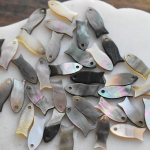 May include: A collection of small, iridescent fish-shaped shell beads in various shades of gray, black, and cream. The beads are scattered on a white, organic-shaped surface, creating a textured and visually appealing arrangement. The fish have a small hole near the head.