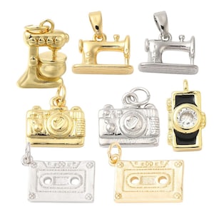 May include: A collection of gold and silver-toned charms. The charms include a stand mixer, sewing machine, camera, and cassette tape. One charm features a black and gold design with a clear stone.