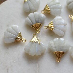 May include: A collection of white moonstone shell charms with gold accents. The charms are shaped like a fan and have a hole at the top for attaching to jewelry.
