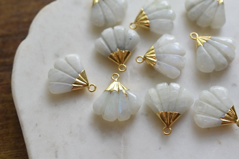May include: A collection of white moonstone shell charms with gold accents. The charms are shaped like a fan and have a hole at the top for attaching to jewelry.