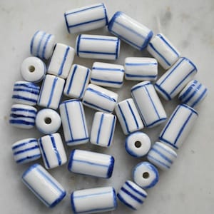 May include: A collection of cylindrical ceramic beads featuring a glossy white finish with hand-painted blue stripes. The beads vary in length and orientation, with some displaying vertical stripes and others showing horizontal bands around the circumference.