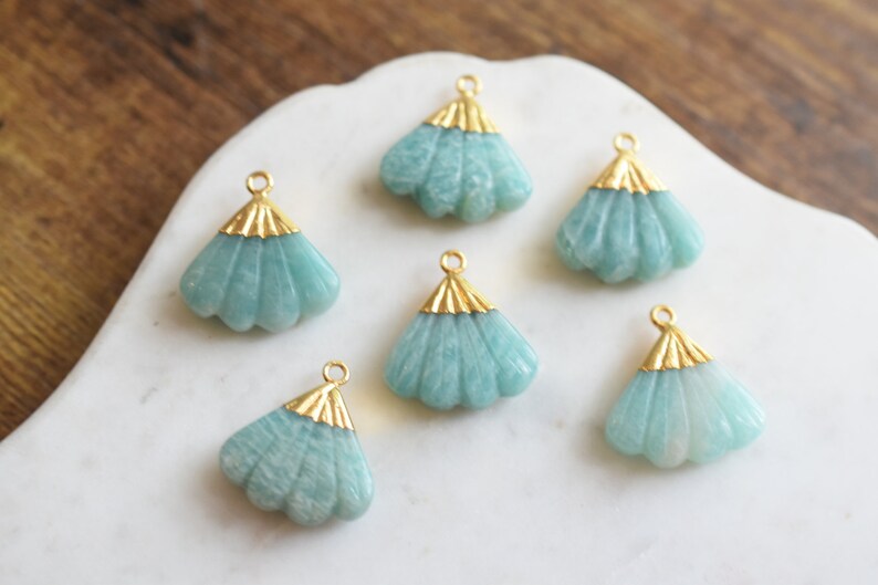 May include: Six teal green gemstone pendants with gold accents in the shape of a fan. The pendants are on a white surface.