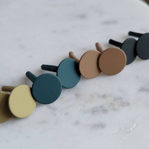 May include: Six pairs of small, round, matte-finish earrings in various colors, including black, brown, green, and beige. The earrings are displayed on a white marble surface.