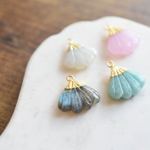 May include: Four gold-toned fan-shaped pendants with different colored stones. The pendants are on a white marble surface. The stones are white, pink, green, and blue.