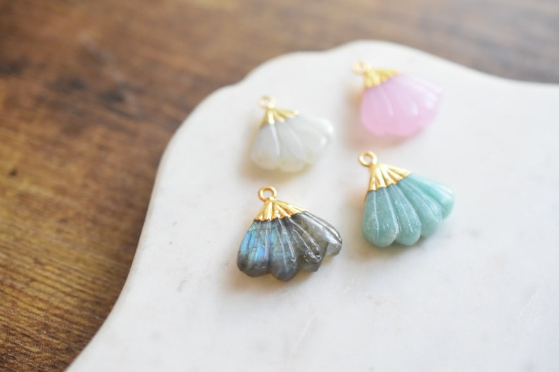 May include: Four gold-toned fan-shaped pendants with different colored stones. The pendants are on a white marble surface. The stones are white, pink, green, and blue.