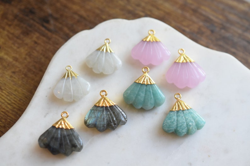 May include: Eight gold-plated shell-shaped charms with different colored stones. The charms are arranged on a white surface. The stones include white, pink, green, and gray.
