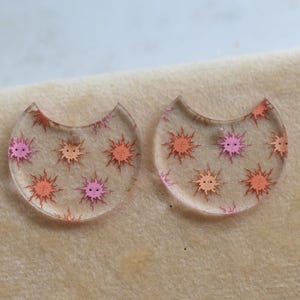 May include: Two semi-circular, clear acrylic earring components featuring a repeating pattern of small, smiling suns in shades of pink and orange. Each piece has a small hole at the top for attachment, showcasing a flat, smooth surface design.