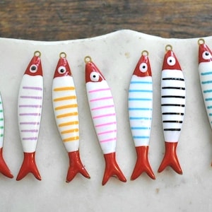 May include: Seven colorful fish-shaped charms with red tails and heads, white bodies, and striped patterns in various colors. Each charm has a gold-colored loop at the top for hanging. The charms are arranged on a white, textured surface.