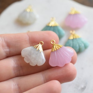 May include: Two gold-tone fan-shaped pendants with a white and pink stone. The pendants are held in a hand.