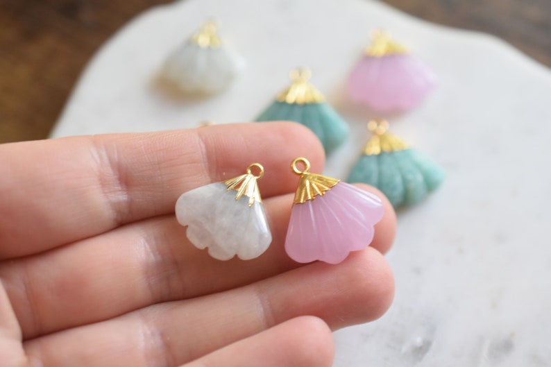 May include: Two gold-tone fan-shaped pendants with a white and pink stone. The pendants are held in a hand.