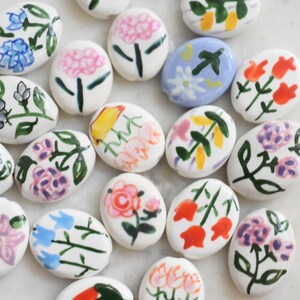 May include: A collection of oval-shaped ceramic beads, each featuring hand-painted floral and fruit designs in various colors including pink, blue, orange, and green on white or light blue backgrounds, arranged in a scattered, loose composition.