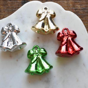Puede incluir: Cuatro colgantes de campana navideña en plata, oro, rojo y verde. Cada campana tiene un lazo en la parte superior y un lazo para colgar. Los colgantes están sobre una superficie blanca, similar al mármol, con un fondo de madera.