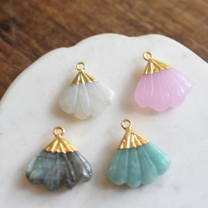 May include: Four gold-tone fan-shaped pendants with different colored stones. The pendants are on a white surface. The stones are white, pink, blue-green, and gray.