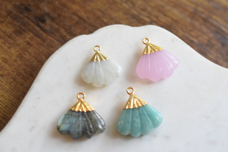 May include: Four gold-tone fan-shaped pendants with different colored stones. The pendants are on a white surface. The stones are white, pink, blue-green, and gray.
