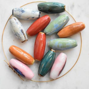 May include: A collection of twelve elongated, cylindrical ceramic beads in various colors including white with blue speckles, solid orange, light green, dark teal, and pink with dark accents, all arranged on a thin, circular gold-toned wire hoop.