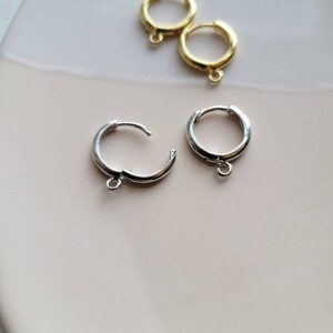 Puede incluir: Tres hallazgos de pendientes de aro, dos dorados y uno plateado. Los hallazgos est&aacute;n sobre una superficie blanca y rosa.