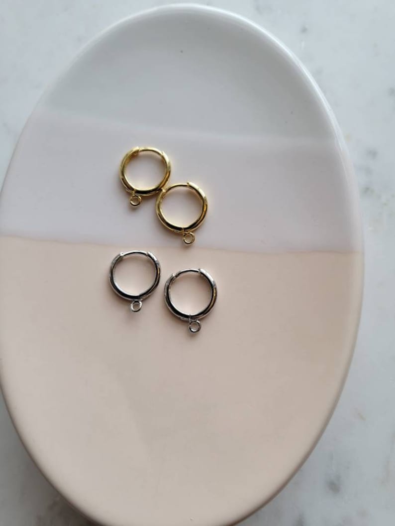 Puede incluir: Dos pares de pendientes de aro, un par es dorado y el otro par es plateado. Los pendientes est&aacute;n en un plato blanco y rosa.