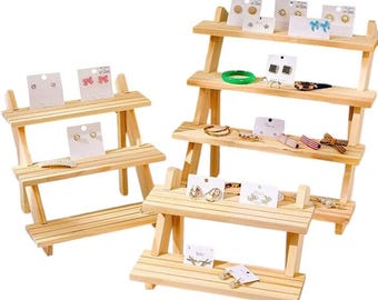 Puede incluir: Expositores de madera con múltiples niveles que muestran pendientes y otras joyas. Los pendientes se presentan en tarjetas blancas. Las joyas incluyen pulseras y anillos. Los expositores están hechos de madera clara.