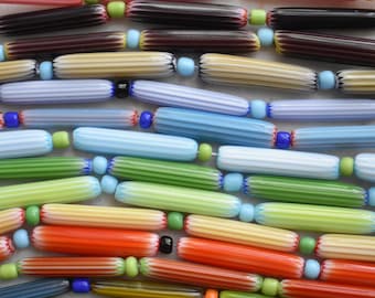 Puede incluir: Una colección de cuentas de vidrio multicolor dispuestas en filas horizontales. Las cuentas son cilíndricas con una textura acanalada y presentan varios colores, incluyendo naranja, rosa, negro, crema, azul, verde y amarillo, separadas por pequeñas cuentas redondas.